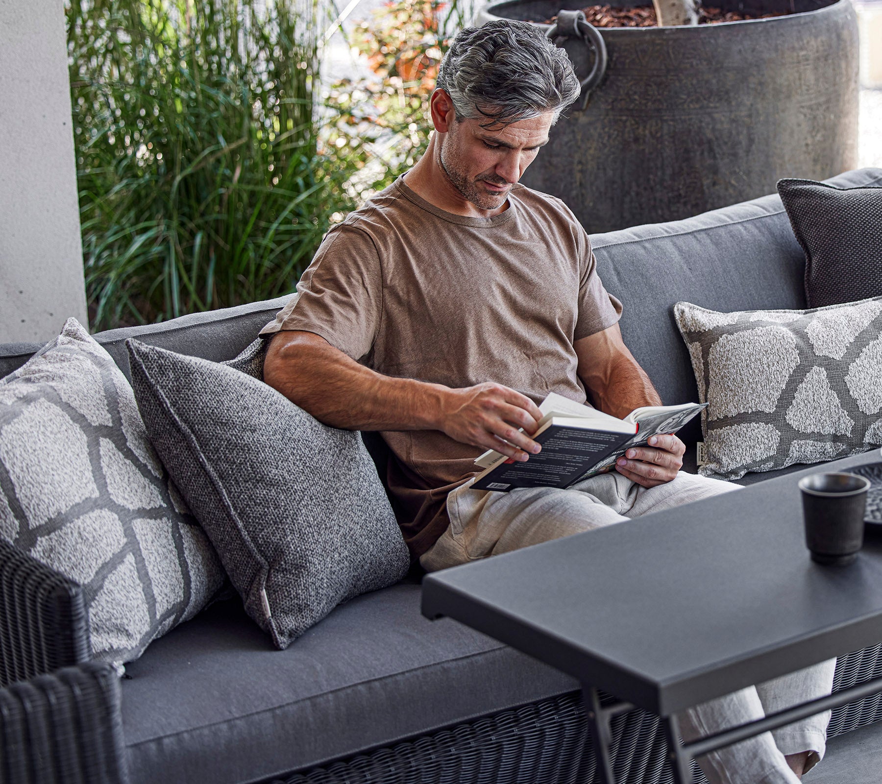 A man reads a book while seated on a cozy sofa adorned with decorative pillows, with greenery in the background.