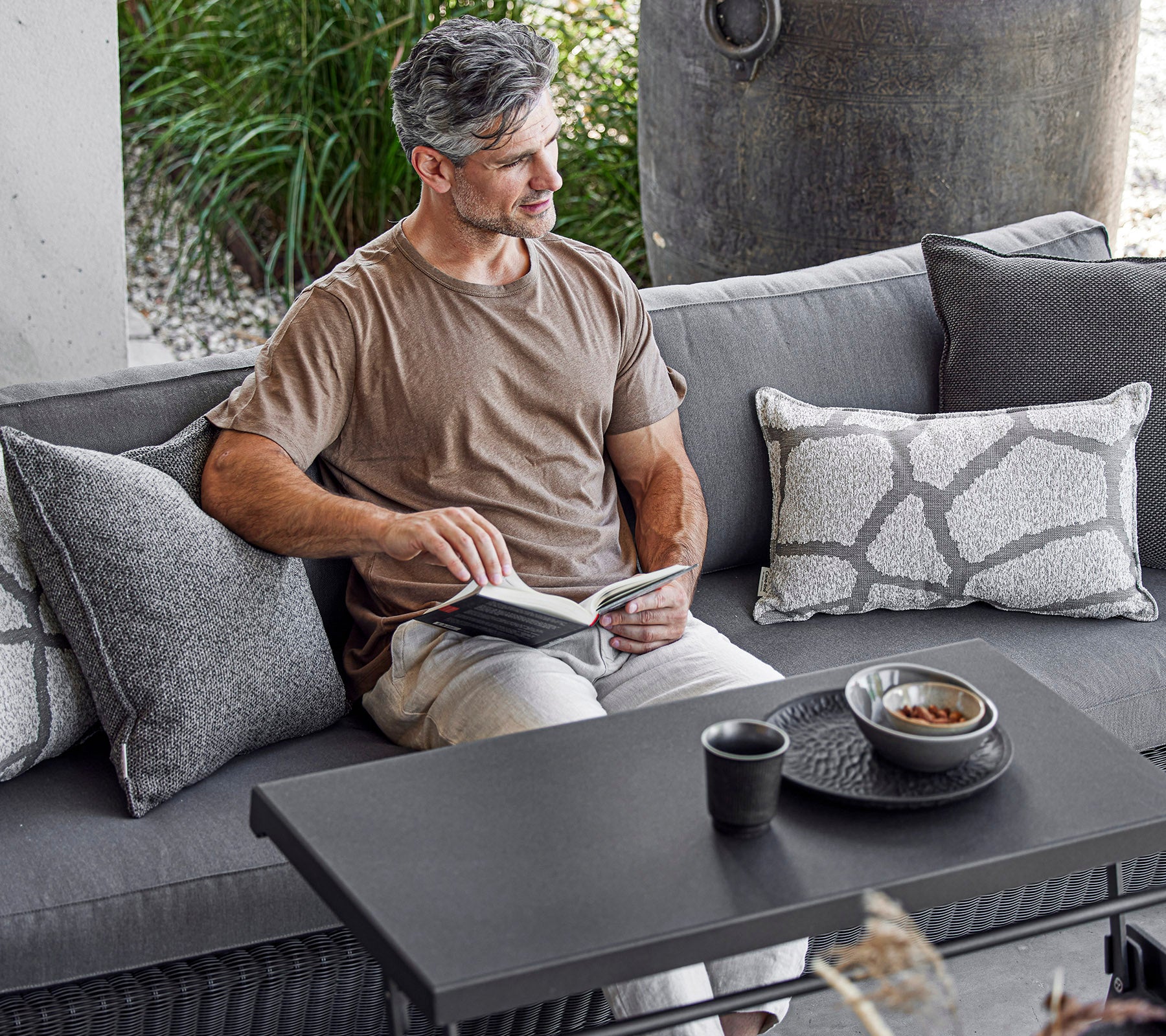 A man sits comfortably on a sofa, reading a book with a cup and a bowl on a coffee table beside him, surrounded by decorative cushions.