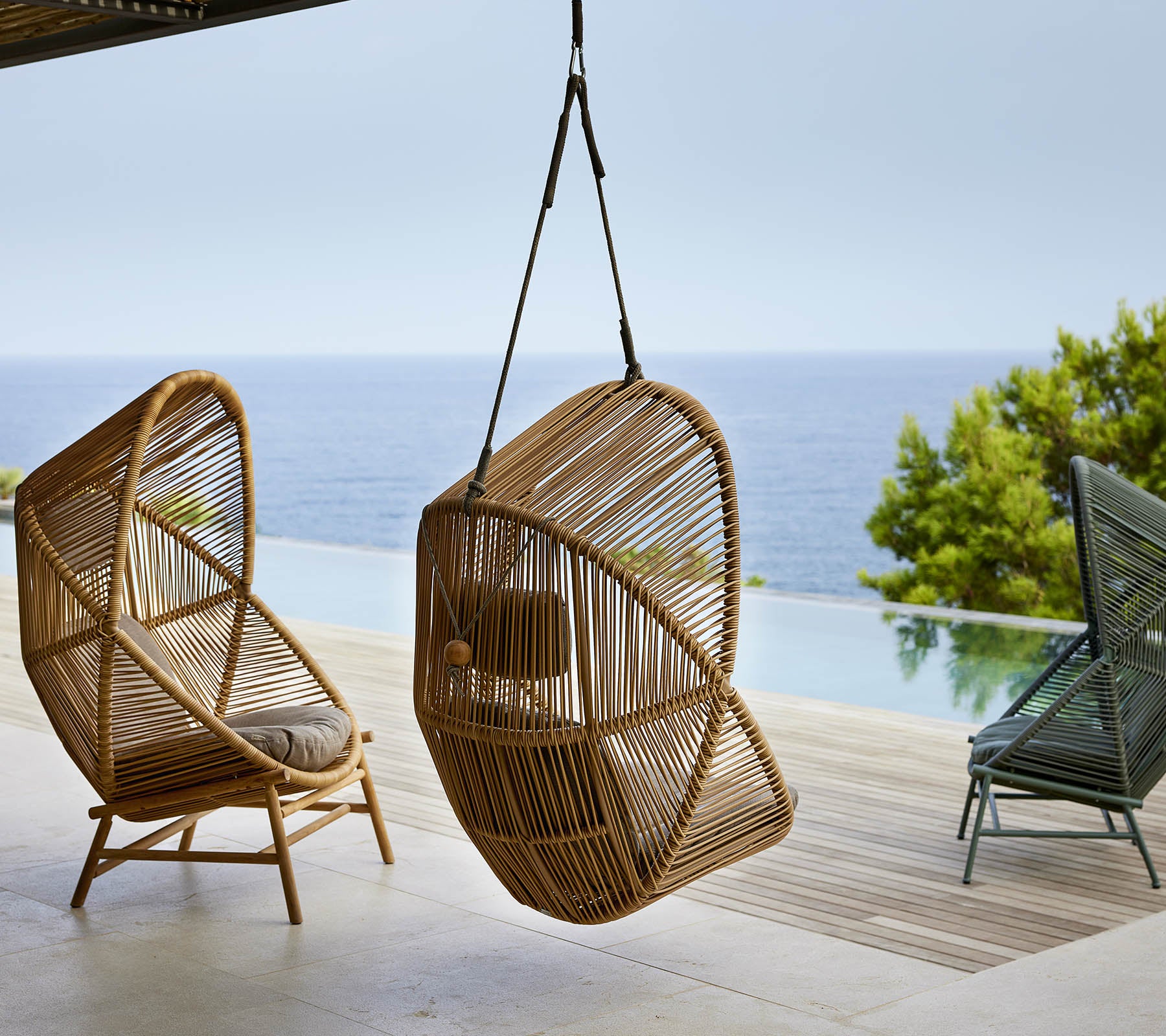 Pair of stylish hanging chairs made of woven material, positioned near a serene body of water, with greenery in the background.
