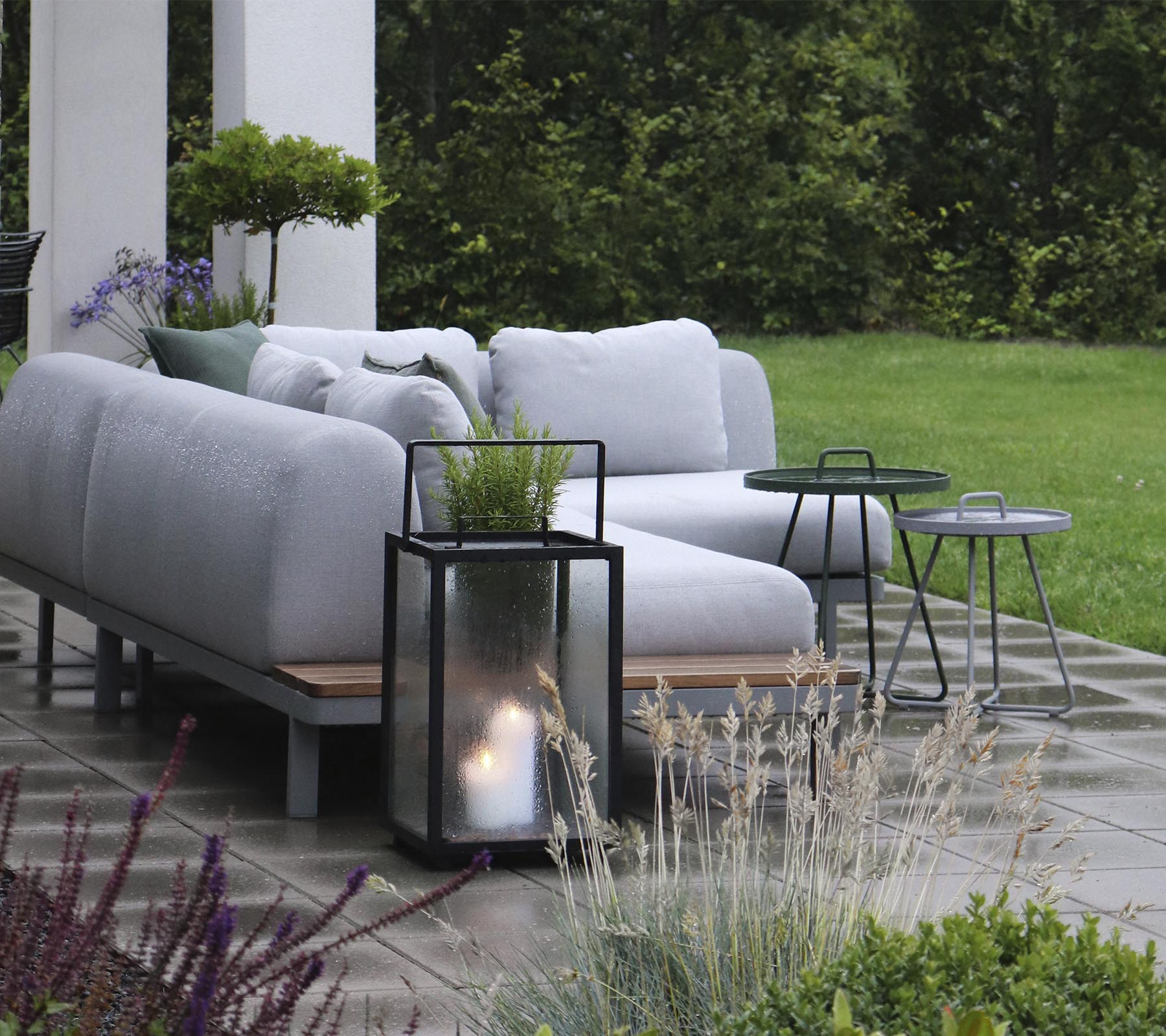 Stylish outdoor seating arrangement with plush cushions, accompanied by small side tables and a lantern, surrounded by greenery.