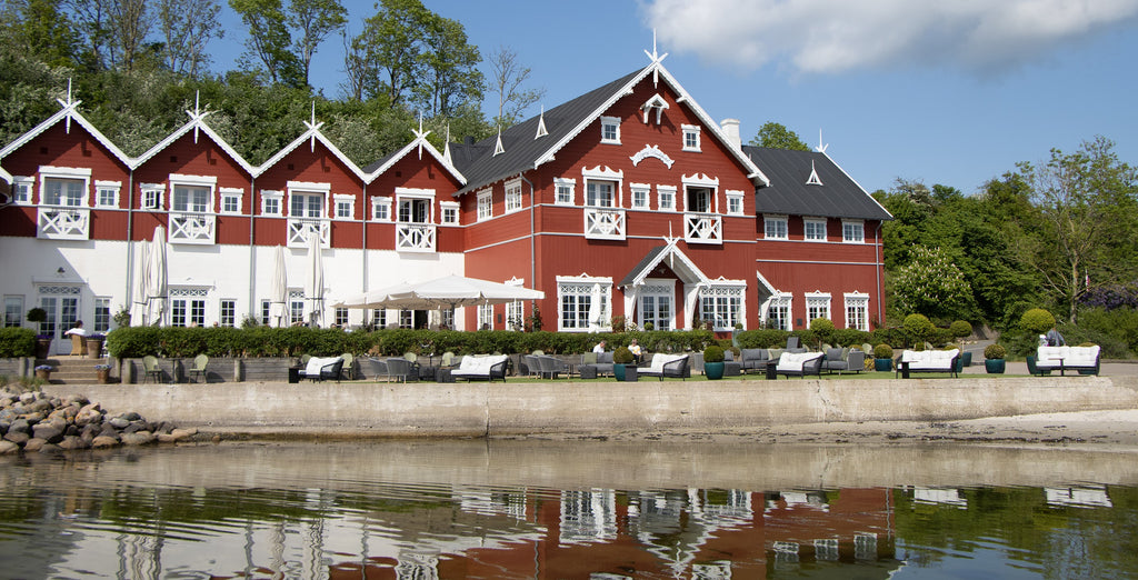 The view of Dyvig Badehotel in Denmark. A red well kept building with white exteriors from the 18th century. Amodern outdoor lounge with lounges sofas in grey and black from Cane-line with ocean view. 
