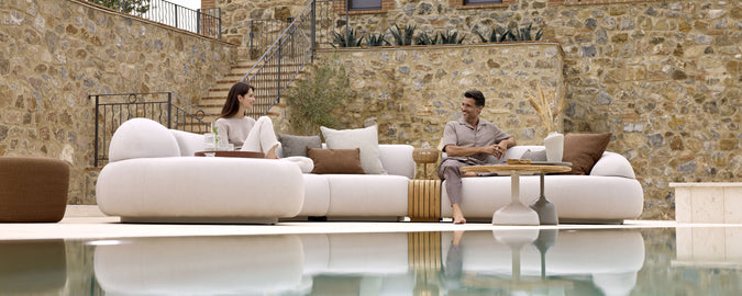 Two people relax on a stylish, modern sofa with cushions, sharing a conversation by a serene water feature and a stone wall.