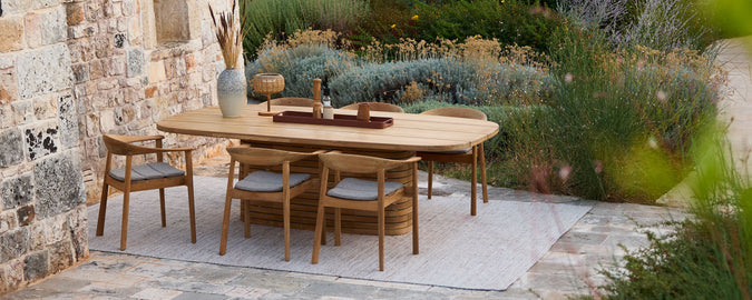 Wooden dining table with wooden chairs and grey cushions in an outdoor setting.