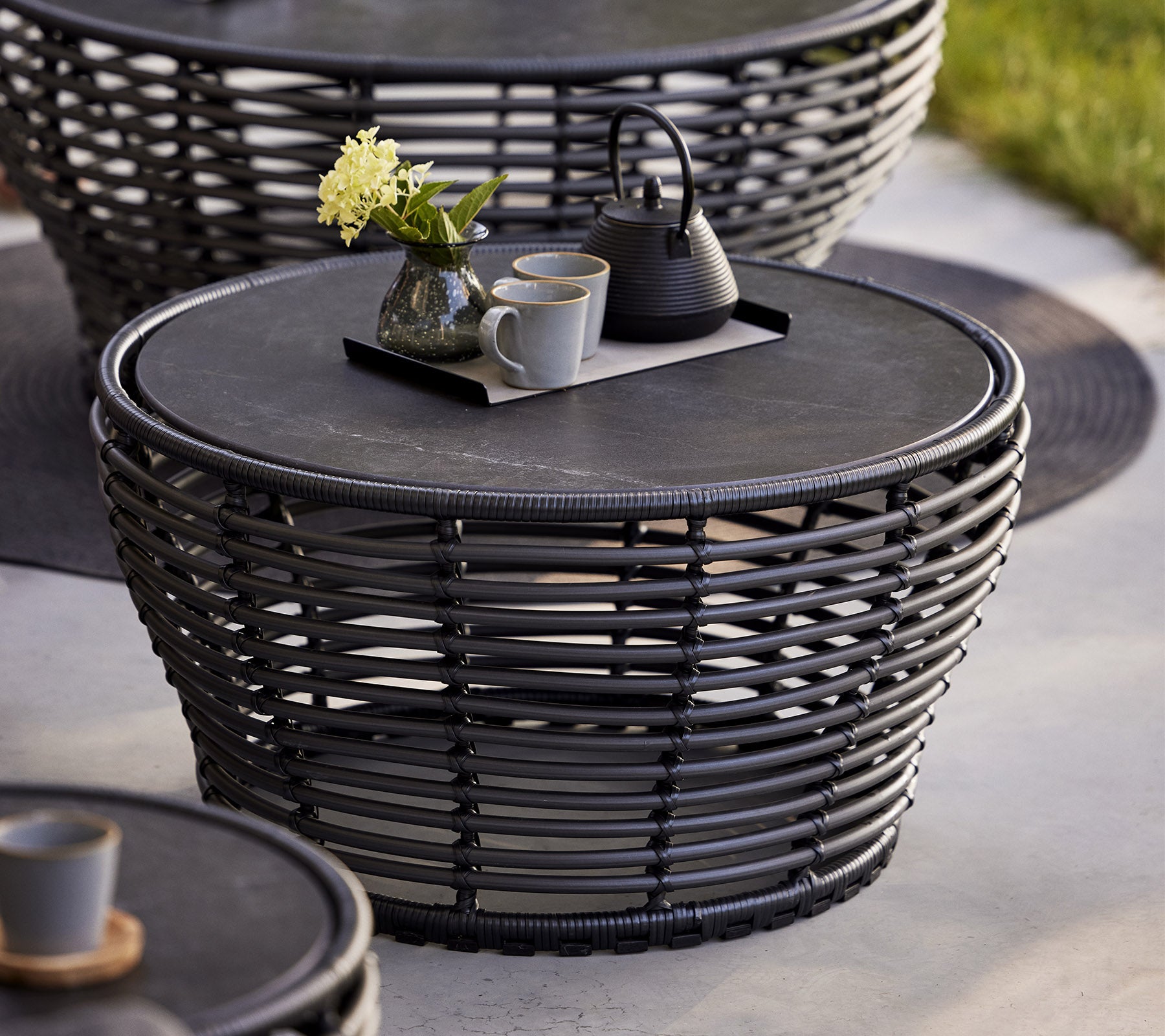 Stylish woven coffee table with a round top, featuring a decorative tray with a teapot and cups, complemented by a flower vase.