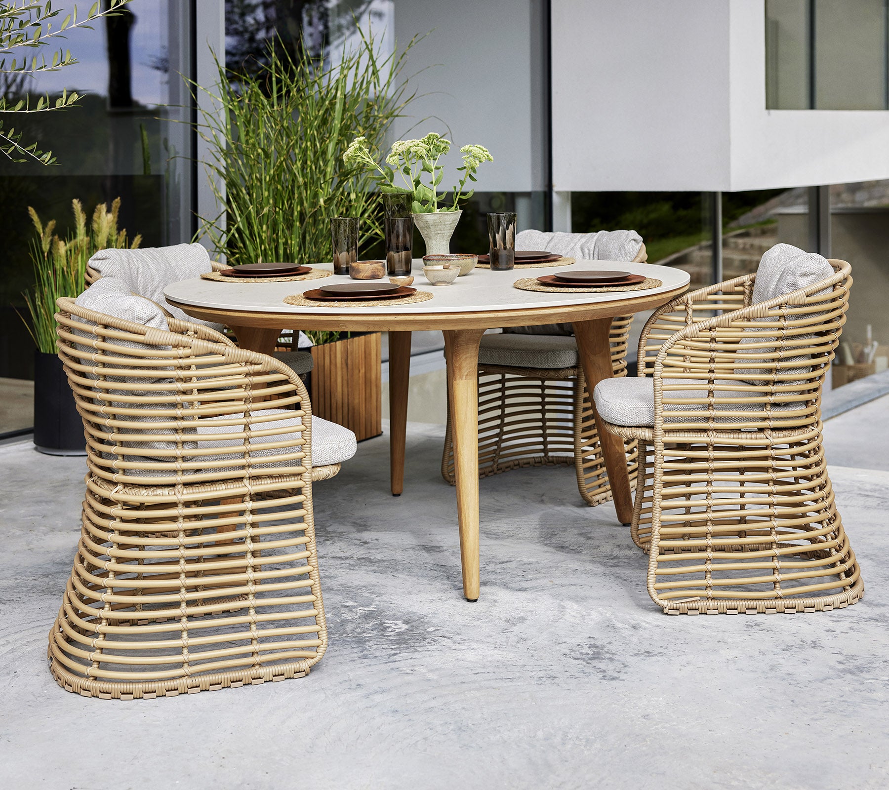 A round table set with four wicker-style chairs, featuring cushions, surrounded by greenery for a stylish dining area.