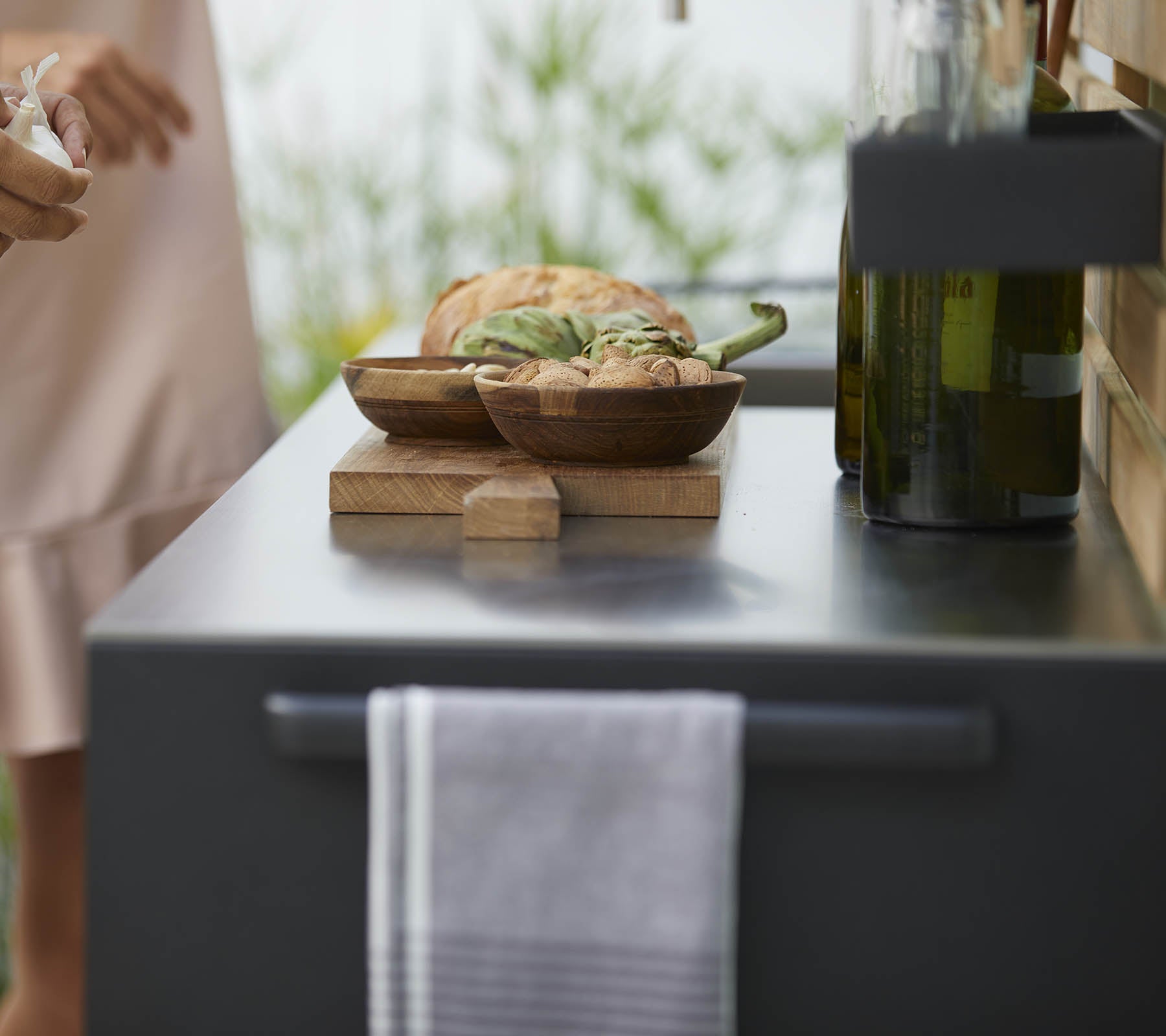 A close-up of a kitchen counter with wooden bowls of food, a towel, and olive oil bottles, with soft greenery in the background.
