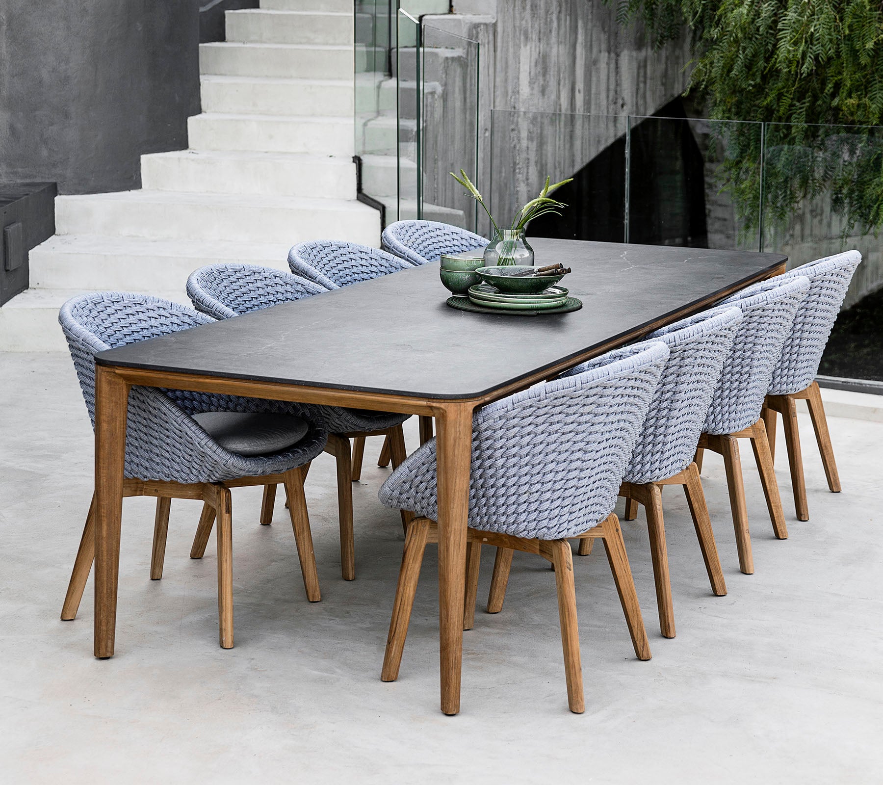 A stylish dining table with a dark surface, accompanied by woven chairs in a light color, complemented by decorative plates and foliage.