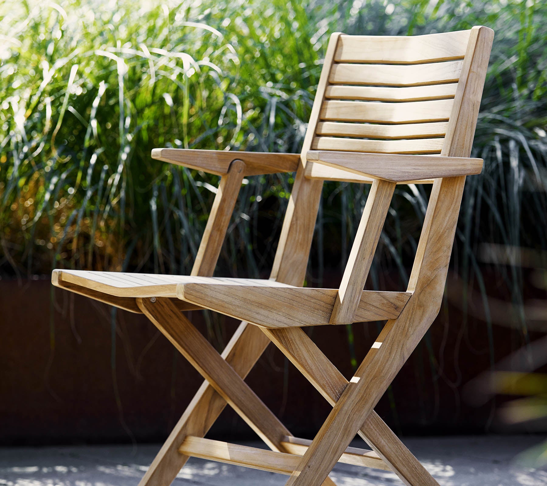 A wooden folding chair with slatted details and armrests, surrounded by lush green foliage. Ideal for relaxation and outdoor settings.