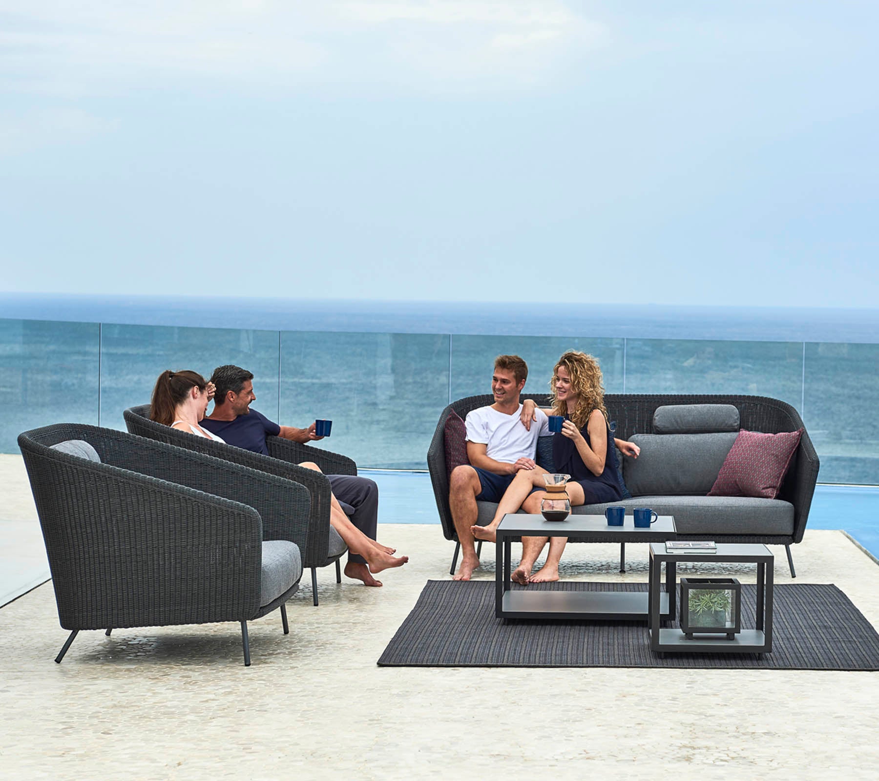 A group of four people socializing on modern outdoor furniture with a view of the ocean, enjoying drinks and conversation.
