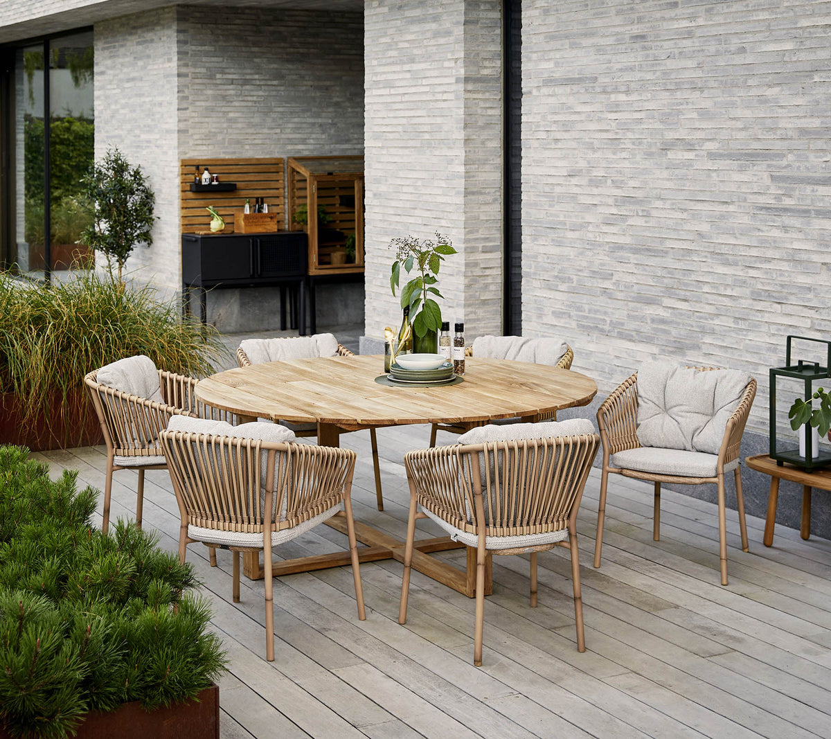 Round wooden dining table surrounded by upholstered chairs, with a decorative plant centerpiece, set against a textured stone wall.