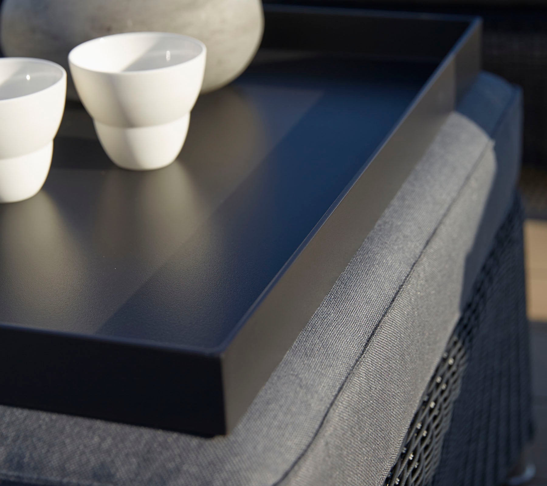 A black tray holds two white cups and a smooth stone, positioned on a textured surface. Soft lighting enhances the scene.