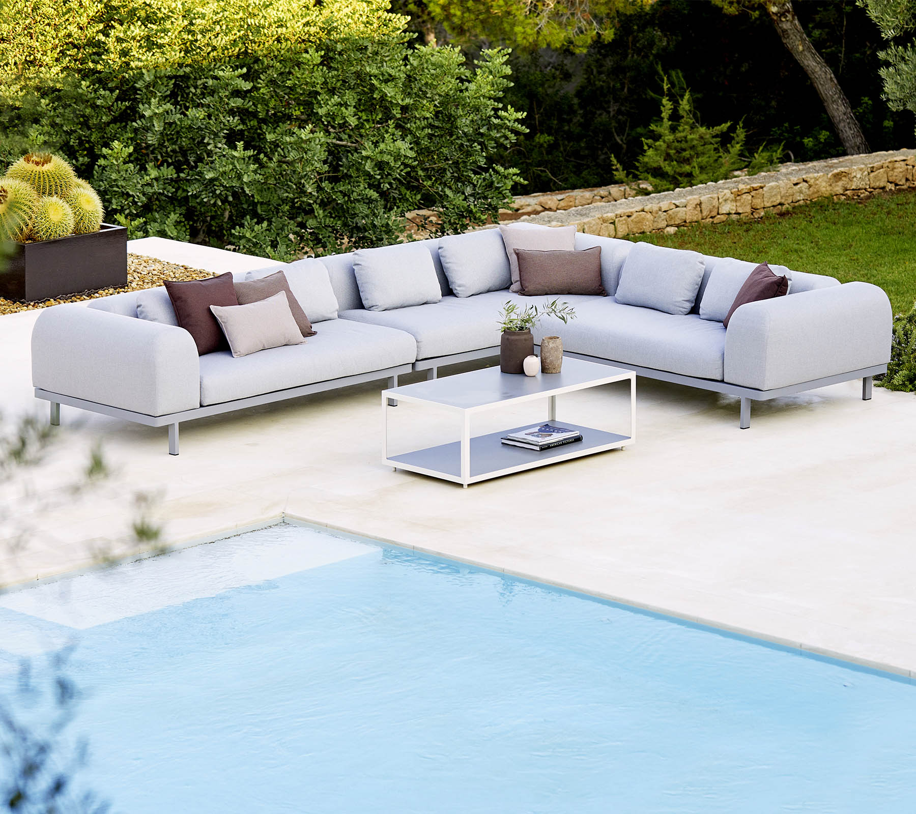 Light grey sectional sofa with decorative cushions and a glass coffee table, positioned next to a swimming pool and lush greenery.