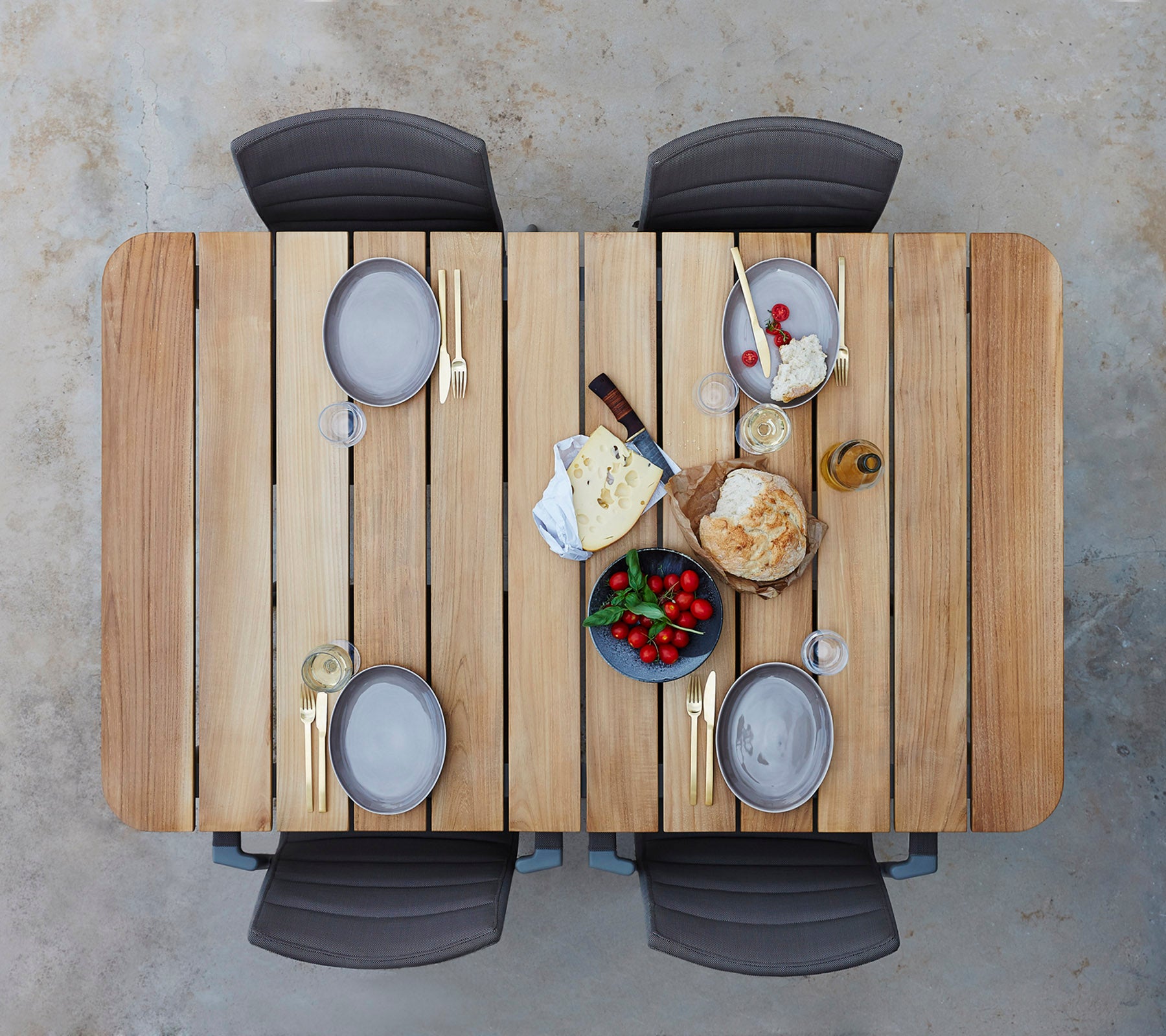 A wooden table set with plates, glasses, and an assortment of cheeses, fruits, and bread, surrounded by four chairs.