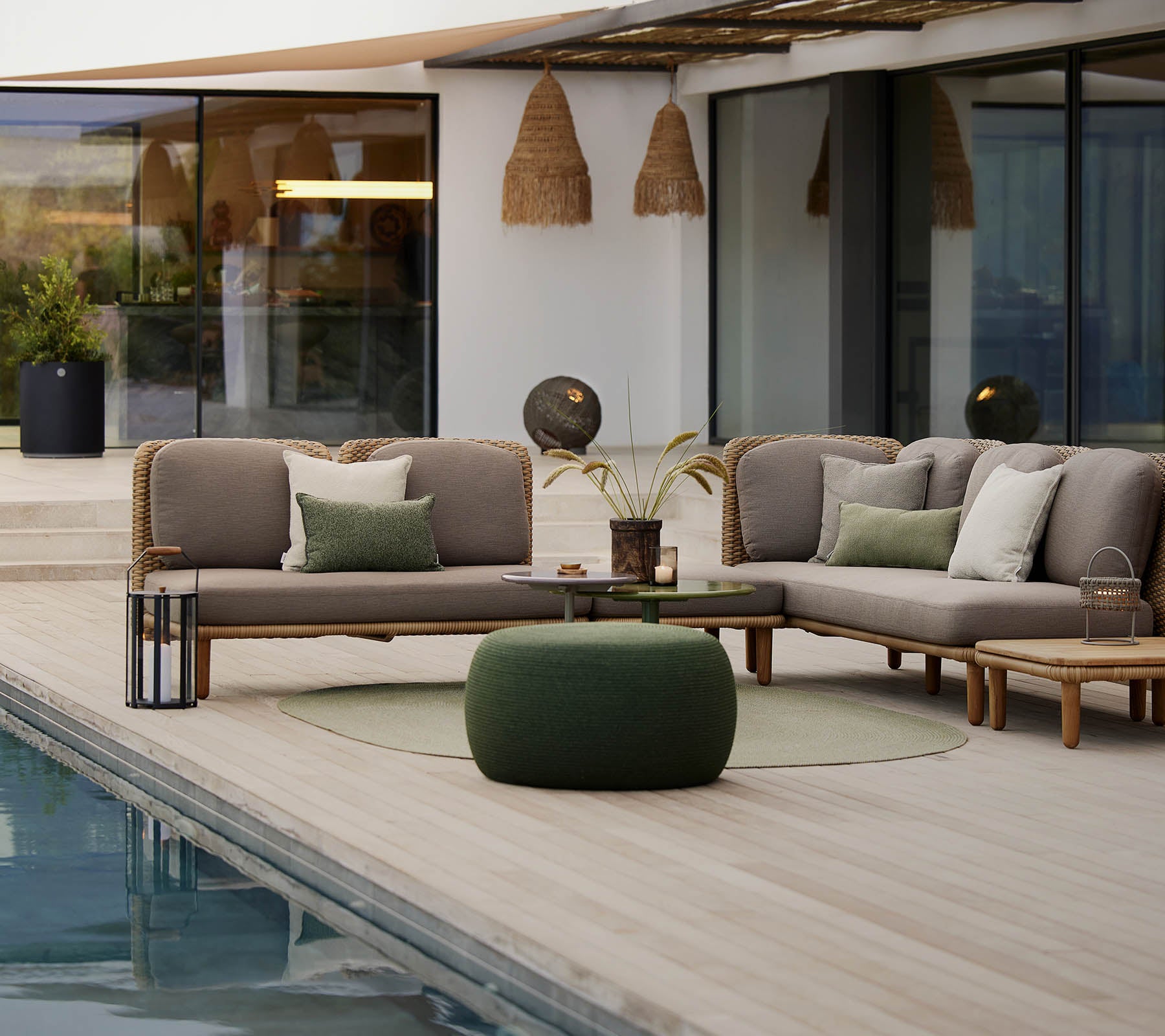 A stylish lounge area featuring a sectional sofa with cushions, a round table, and a cozy pouf, set beside a tranquil pool.