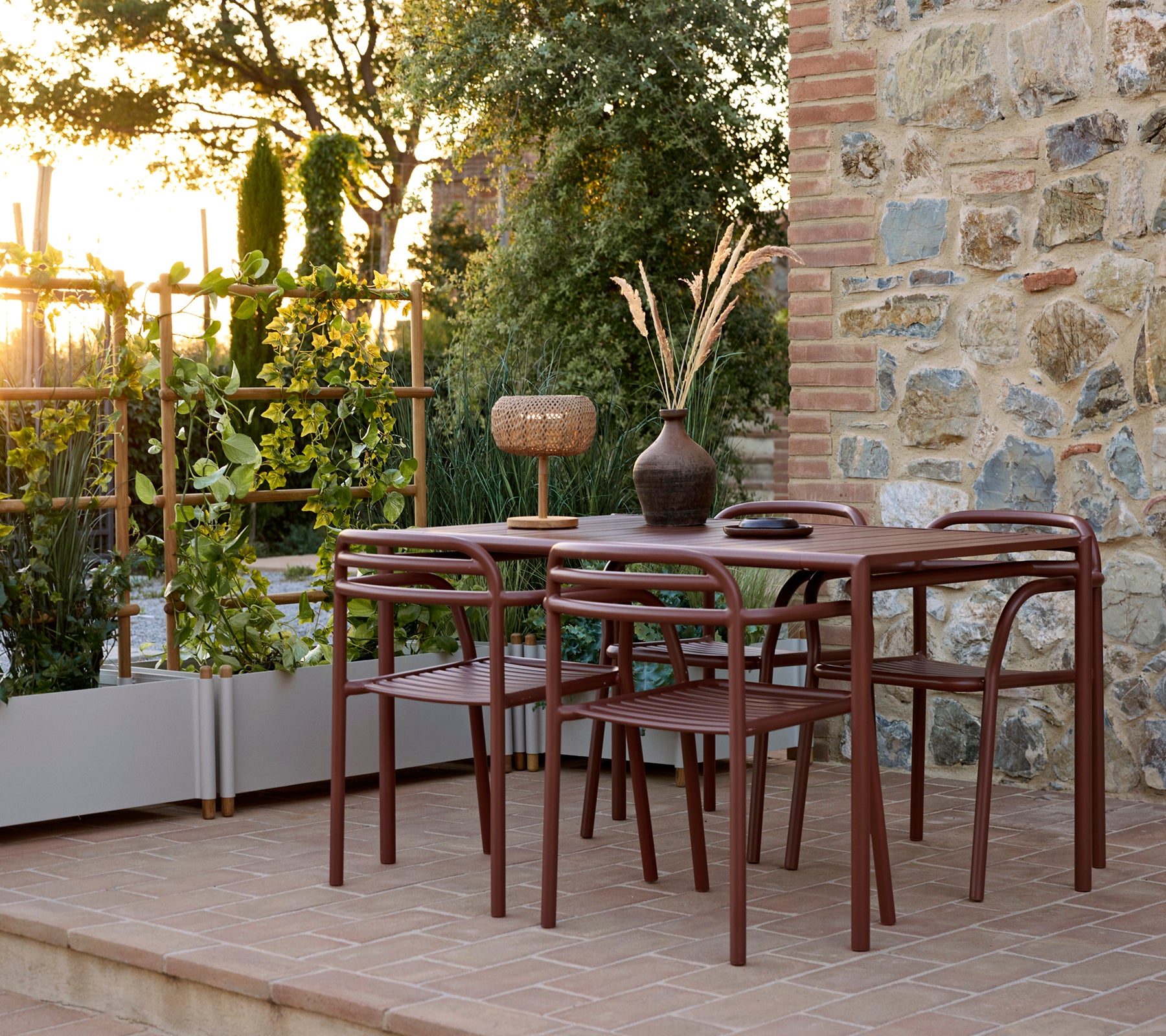 Outdoor setting featuring a Bliss table with chairs, a Bliss armchair, and a Sticks planter surrounded by greenery at sunset.