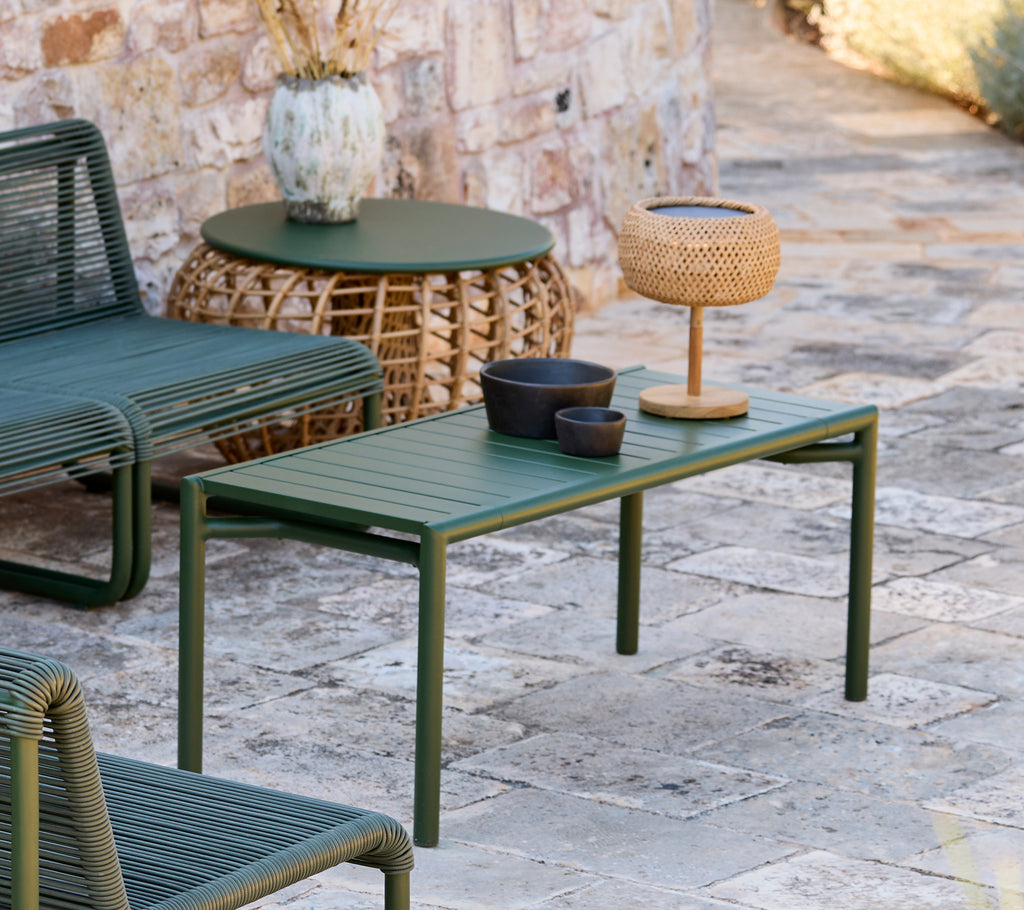 Green table with black bowl, brown vase, and woven lamp in a modern outdoor lounge area.