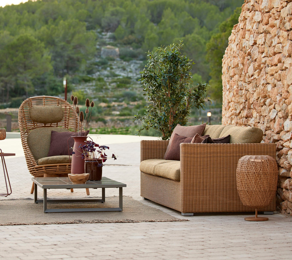 Luxurious outdoor seating area featuring a woven sofa, a stylish chair, a coffee table, and decorative elements surrounded by greenery.