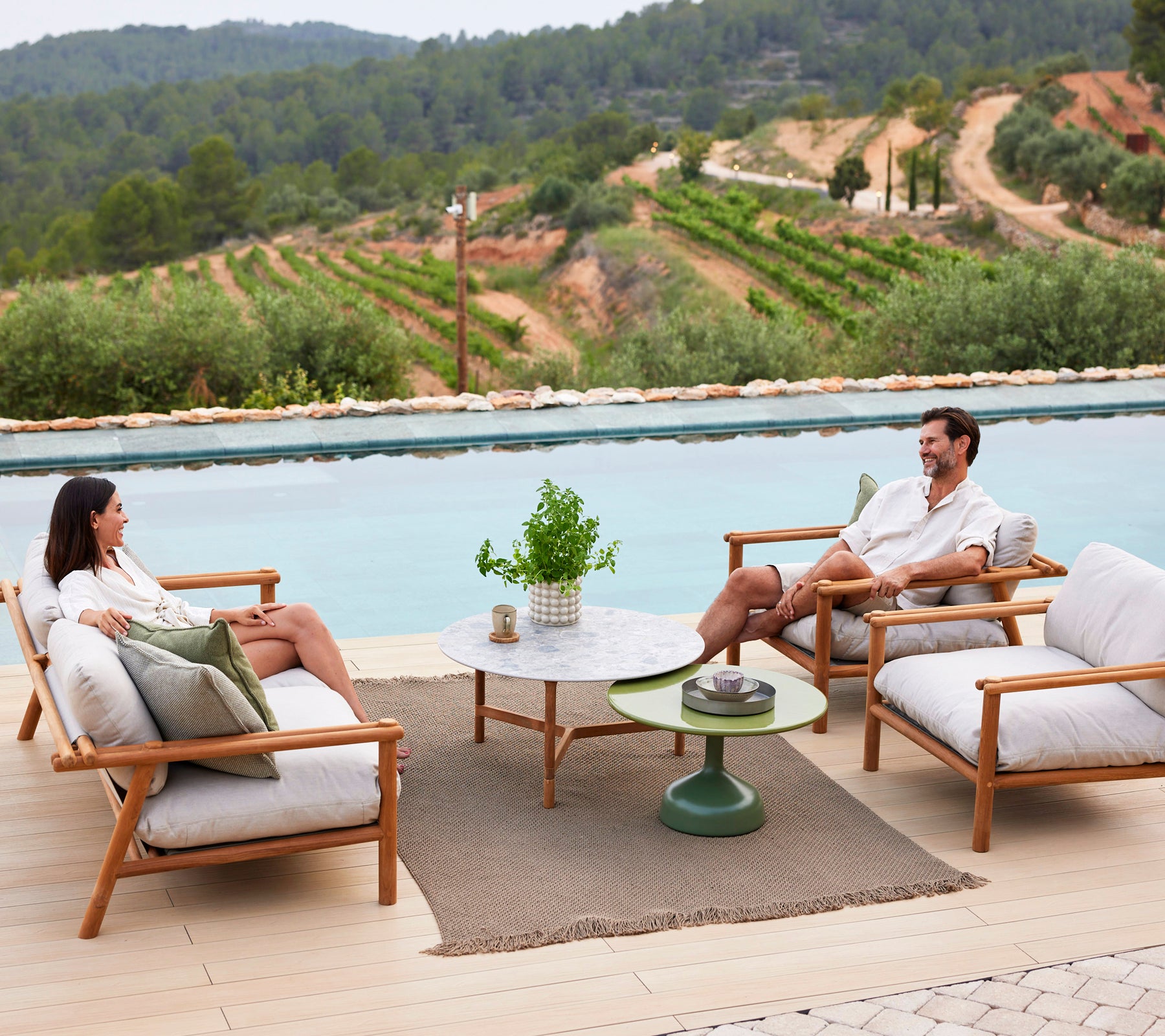Two people relax on lounge chairs around a coffee table, with a knit rug beneath and scenic greenery in the background.