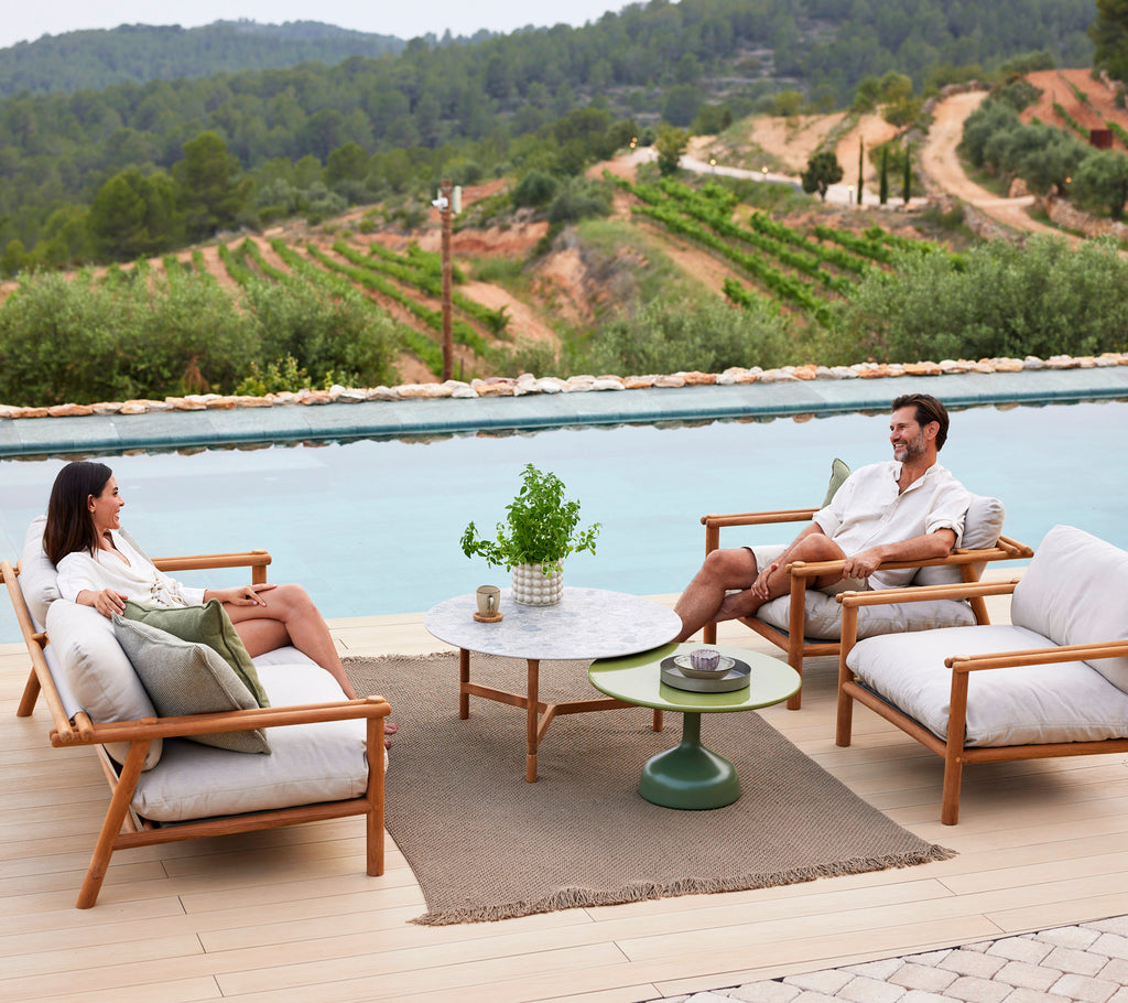 Two people relax on lounge chairs around a coffee table, with a knit rug beneath and scenic greenery in the background.