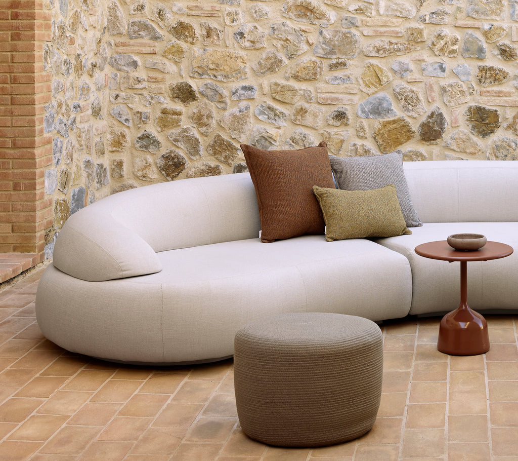 Curved sofa with decorative pillows, a circular footstool, and a glazed side table on a textured floor with a stone wall backdrop.