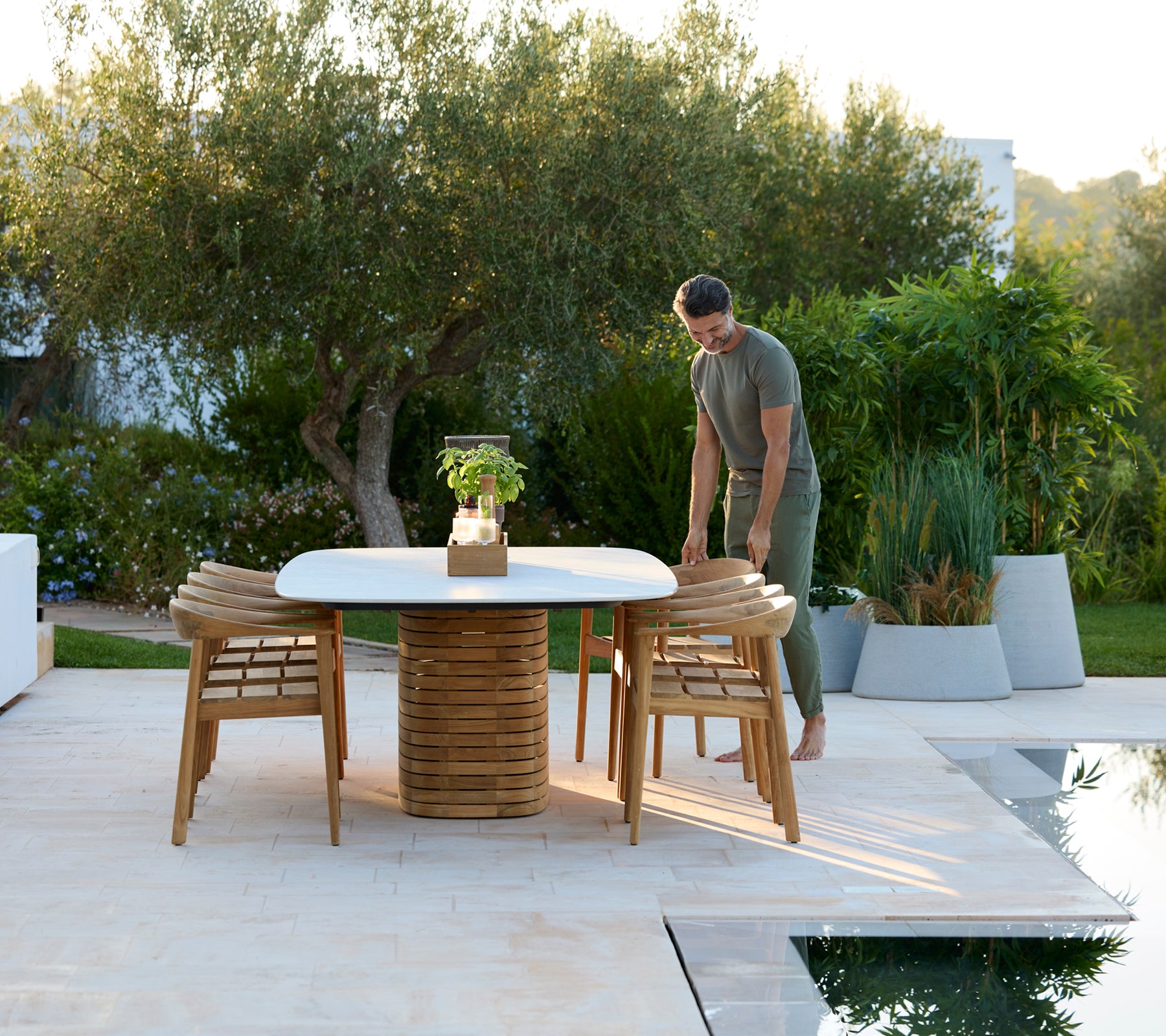 Modern outdoor round table with wooden chairs and greenery in the background.