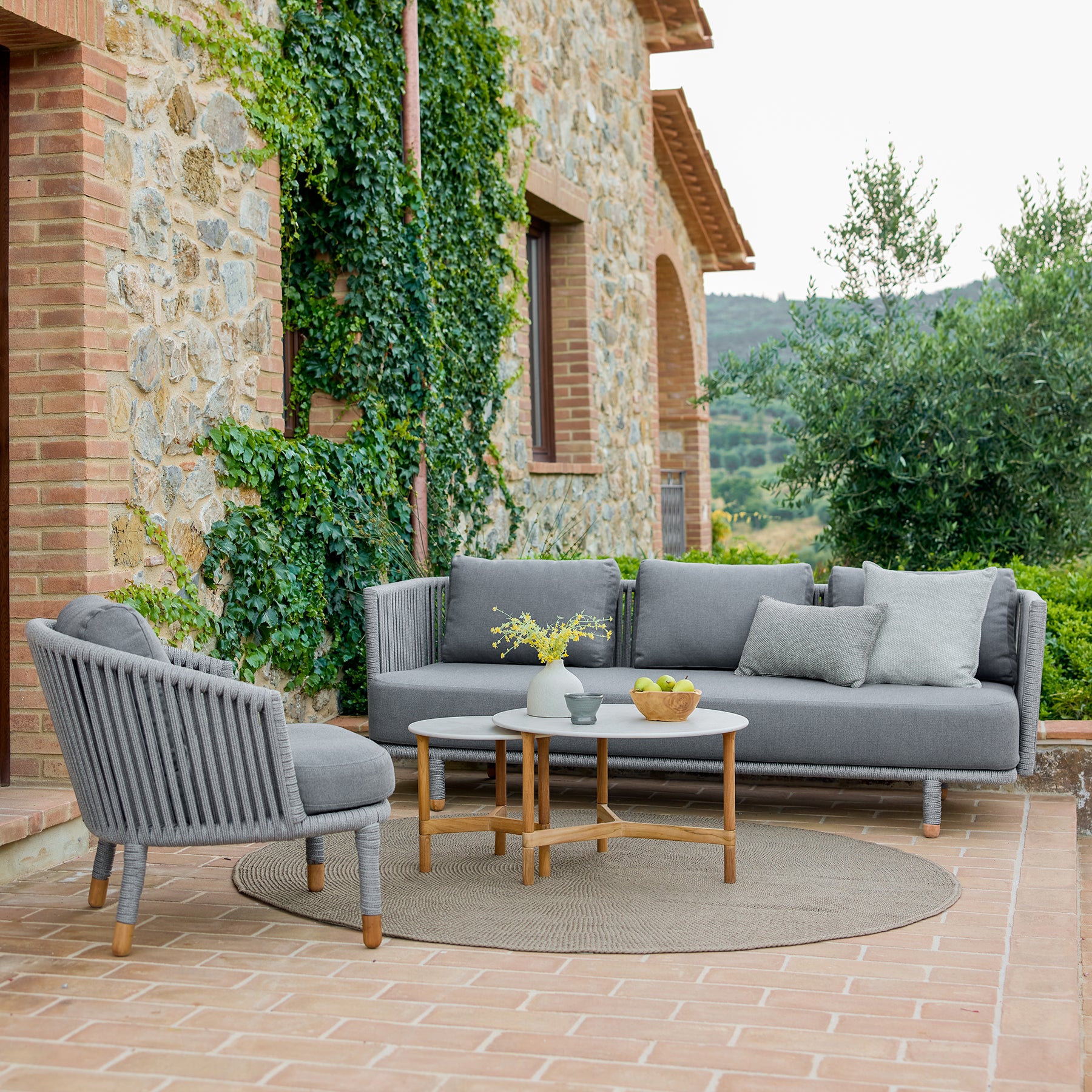Modern grey sofa set with matching armchair and a round coffee table, surrounded by greenery and rustic stone architecture.