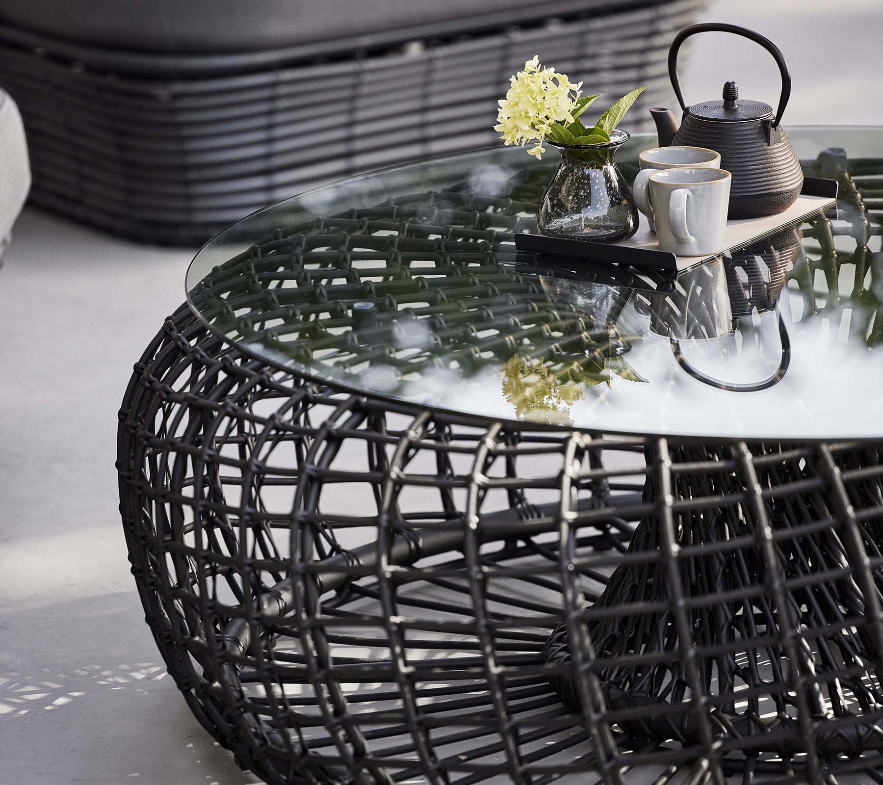 Nest coffee table featuring a woven base, a glass top, and a tea set with flowers for a stylish, modern look.