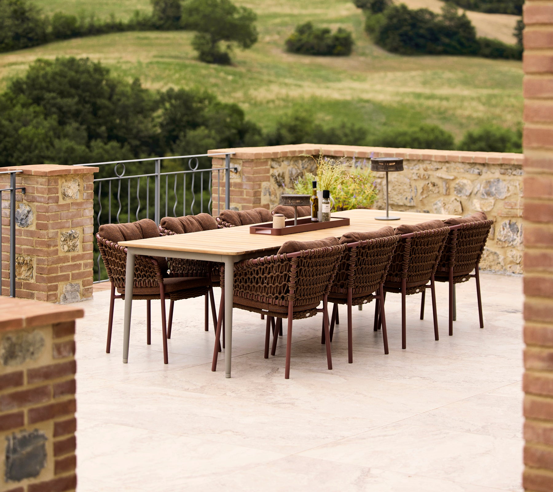 Umber brown Ocean dining chairs surrounding a Core dining table, set against a lush green landscape. Elegant outdoor dining arrangement.