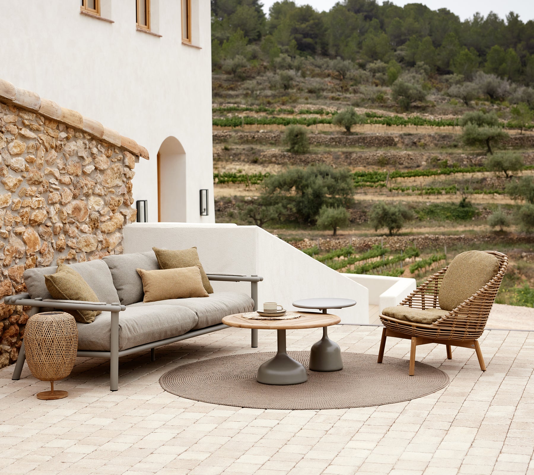 A stylish lounge area featuring a cozy sofa with cushions, a round rug, a modern side table, and a woven lounge chair, surrounded by greenery.